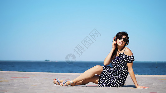 戴着太阳镜和太阳裙 戴着大耳机 在海滩上 在炎热的夏日 靠近海边的智能手机上听音乐的美丽黑发女孩电话青年耳机乐趣旅游幸福闲暇晴天图片