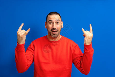 在孤立的蓝色背景上穿着红色套头衫的迷人西班牙裔男子的画像用疯狂的表情尖叫着举起手来做摇滚符号 音乐明星 重概念图片