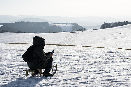 一个男人看着他的手机 冬天坐在雪山顶上的寒冷中 坐在儿童雪橇上 裹着他的冬季保暖夹克享受冒险成就冻结自由思维外套沉思男性荒野图片