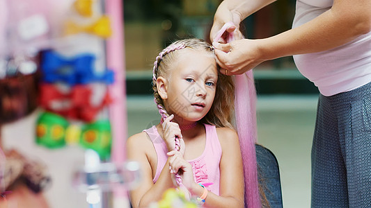 美丽的金发女孩 七岁 编着两条辫子 在美容院 美发沙龙的大镜子前用粉红色的头发做发型 一个小公主黑发造型师父母职业成人女士编织治图片
