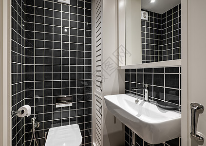 漂亮的浴室配有黑色光滑的方形瓷砖 带水龙头的白色陶瓷水槽和木制毛巾储物柜 镜子映出天花板房间的上半部分 有灯和通风图片