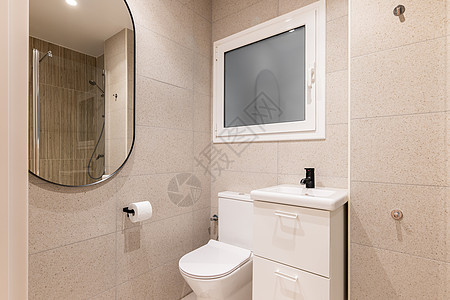 带马桶的浴室的一部分 桌子上的洗脸盆 墙上漂亮的椭圆形镜子 内衬米色大理石瓷砖 您可以在镜子中看到带金属配件的淋浴区的倒影洗手间图片