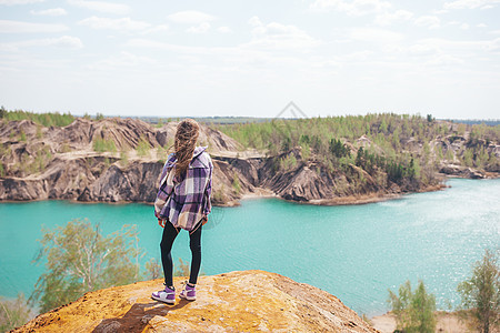 美丽的女孩在岩石的顶端 有蓝湖背景背景上悬崖活动石头蓝色海湾娱乐远足者闲暇远足游客图片