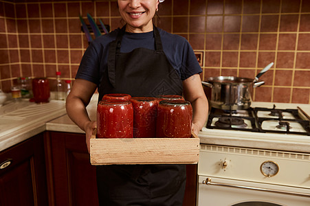穿着黑色厨师围裙的可爱女性家庭主妇 拿着一个木箱 装满罐子番茄糊和酱汁倒在地上图片
