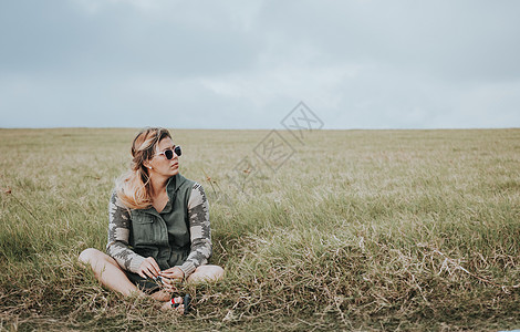 尼加拉瓜戴眼镜的女孩的画像盘腿坐在田里 坐在领域的轻松的青少年的女孩看对边 年轻女性在田间坐着放松的概念假期太阳镜活动绿色休闲场地福利农背景