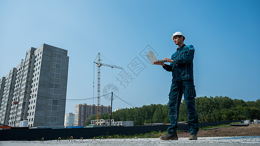 一名戴头盔的男性建筑师站在一个建筑工地的背景之下 手里拿着一台笔记本电脑基础设施工程师建设者项目机械安全帽衣服企业家起重机劳动者图片