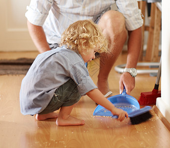 他是一个可爱的清洁工 一个幼儿蹲下扫沙 在他父亲的监管下在背景里图片