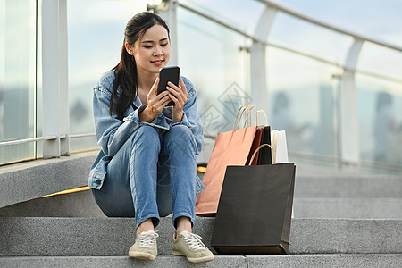 使用移动电话 坐在屋顶楼梯上用袋袋购物时使用手机的充满欢乐的亚洲妇女图片