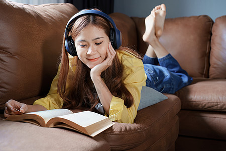 在沙发上、看书亚裔妇女在家里沙发上放松时 阅读书本并戴耳机技术幸福微笑视频女性商务摄影学生音乐成人背景