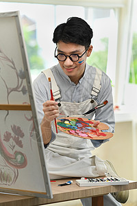 快乐的亚裔男性艺术家持有画笔和调色板绘画 张贴在艺术工作室的画布上活动刷子爱好公寓天赋休闲油画艺术品想像力家庭生活图片