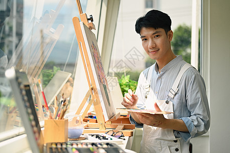 快乐的亚裔男性艺术家持有画笔和调色板绘画 张贴在艺术工作室的画布上杰作艺术品客厅专注爱好油画家庭生活职业活动画家图片