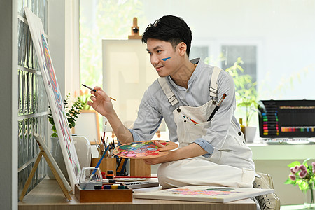 快乐的亚裔男性艺术家持有画笔和调色板绘画 张贴在艺术工作室的画布上作坊职场水彩创造力学生画家公寓客厅爱好家庭生活图片