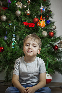 照片上是一个穿着灰色 T 恤的漂亮男孩在圣诞树前看着镜头 在明亮的房间里的肖像 自然的 而不是舞台摄影头发玩具孩子成人白色童年年图片