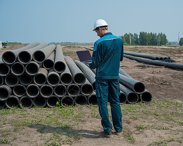 白种男性建筑工身戴硬帽 站在管道附近 在一个建筑工地使用笔记本电脑起重机建设者工厂工人安全帽职场技术安全员工领班图片