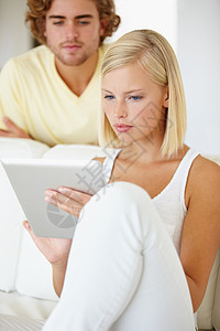 只是偷看一下 一位美丽的年轻女子正在使用平板电脑 而她的男友则在她的肩膀上观看背景图片