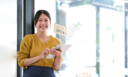 快乐的亚洲年轻女主人 站着拿着一块有开放标志板的平板板生意人士工人咖啡馆女士服务员微笑企业家药片围裙图片
