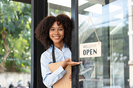 小企业非洲女老板面带微笑 同时转向咖啡馆开业的标志 穿着围裙的快乐的美国黑人女服务员企业家在门上签名成人员工商务女性人士女士职业图片