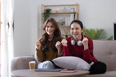 两个年轻的女人在家里的客厅里坐在沙发上看电视 高兴地握着拳头庆祝最喜欢的运动队的胜利 周末休闲 电视节目和娱乐概念享受手表闲暇女图片