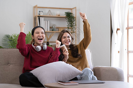 两个年轻的女人在家里的客厅里坐在沙发上看电视 高兴地握着拳头庆祝最喜欢的运动队的胜利 周末休闲 电视节目和娱乐概念微笑喜悦女士长图片