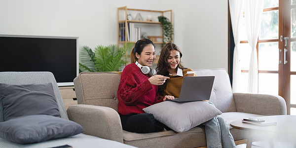 亚洲女性朋友坐在客厅里一起在笔记本电脑上看电影 现代女性友谊在家中享受无线技术的周末活动生活方式长椅电视闲暇爆米花娱乐男性手表家图片