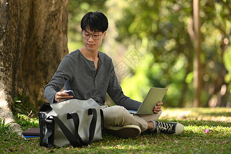 夜间阳光下在树下使用笔记本电脑的年轻亚洲男子学生 教育与生活方式概念图片