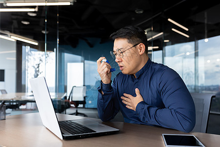 一名在工作场所哮喘病发作的亚洲男子办公室工人 他坐在一张桌子上 使用吸入器图片