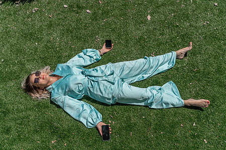 穿着眼镜和蓝色衬衣的月光卷发妇女躺在夏季新鲜绿草上 她手里拿着手机(电话)图片