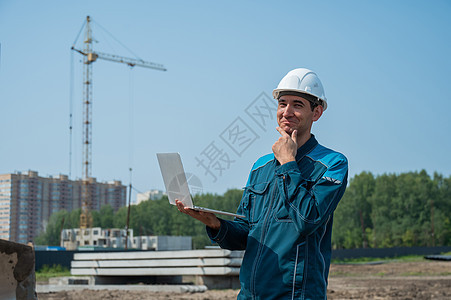 一名戴头盔的男性建筑师站在一个建筑工地的背景之下 手里拿着一台笔记本电脑蓝图基础设施建筑学衣服管道就业领班安全帽检查起重机图片