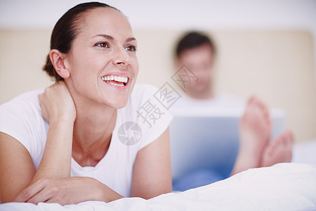 幸福地数着她的祝福 一名年轻女子躺在床上 而她的丈夫在后台使用笔记本电脑工作图片