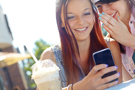 一个十几岁的女孩在手机上发短信 而她的朋友在她耳边悄悄地说着什么话图片