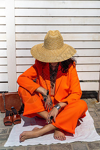 穿着橙色西装 戴帽子的时髦妇女坐在白条纹背景的地毯上 手上有首饰戒指和手镯 凉鞋和一个包袋配件衣服白色旅游橙子奢华自由女性项链女图片