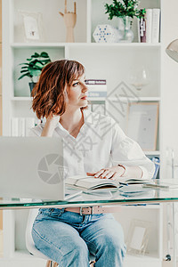 在计算机办公室的女人 她身穿白衬衫和蓝色牛仔裤 坐在桌旁 桌上放着一台笔记本电脑和一个记事本 在办公室工作 明亮的工作场所商务桌图片