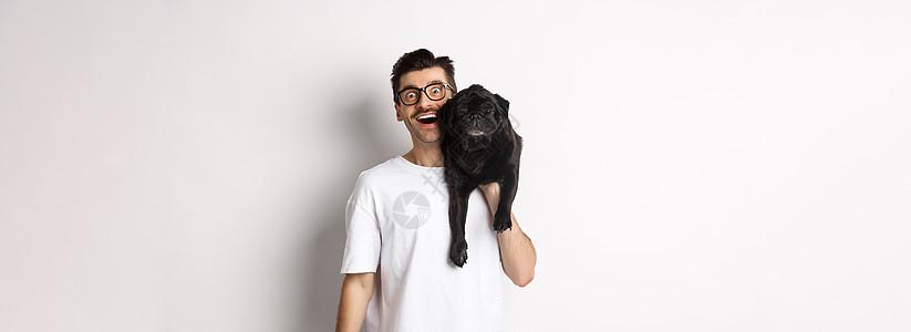 戴着眼镜和 T 恤的快乐嬉皮士 肩上扛着可爱的黑色哈巴狗 面带微笑 狗主人和他的宠物玩耍 站在白色背景上胡须动物促销犬类工作室行图片