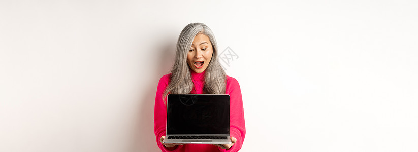 灰色电脑电子商务概念 以灰色头发震惊的亚洲祖母 在网上检查宣传手册 展示笔记本电脑黑屏 站在白背景上站立企业家互联网毛衣时尚快乐技术购物背景