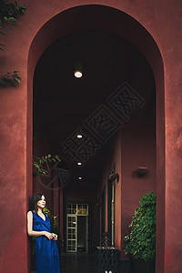 一位身穿蓝色连衣裙的年轻美女站在大拱门下的夏日露台上 靠近一栋赤土色的豪宅图片