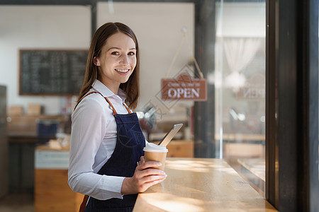 拥有平板牌的餐馆中的年轻女性经理有开放标志的咖啡厅女店主小企业概念服务女士入口围裙施工成功职业店铺药片员工图片