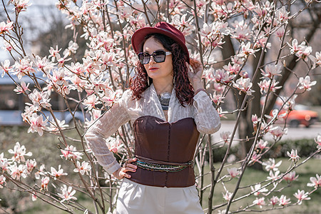 戴帽子的时髦女人站在公园木兰灌木丛附近 穿着白色紧身裤和装着相机 (笑声) (掌声)图片