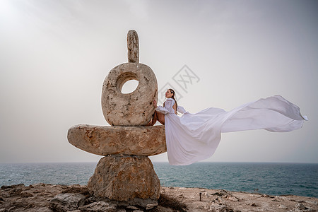 一个女人坐在一块用大石块雕塑的石头上 穿着白色长裙 在海空背景下 风中发芽着洋装和服饰物卵石蓝色冥想旅行岩石地标海洋平衡天空裙子图片