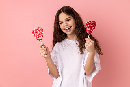 满足的小女孩拿着两颗心形棒棒糖 用快乐的表情看着相机图片