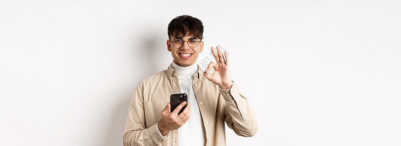 技术和网上购物的概念 戴眼镜的现代帅哥使用智能手机展示 OK 手势 推荐应用程序或商店 白色背景赞扬男人工作室情绪眼镜广告成功学图片