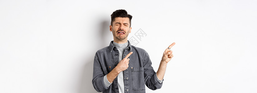 悲哀的哭泣的人用左手指着标志 哭泣和抱怨 嫉妒或沮丧 站在白色背景上夹克胡子工作室快乐苦恼成人男人标识广告男性图片