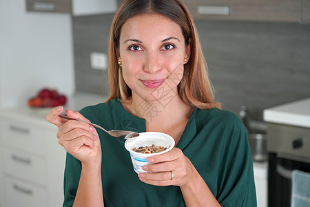 迷人的女人拿着一碗希腊酸奶加面粉 对着摄影机微笑 背景上的厨房模糊不清饮食女士食物勺子美食麦片甜点小吃拉丁粮食图片