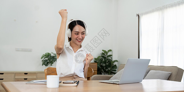 亚洲少女戴着耳机在笔记本电脑上在线学习时听音乐 快乐的女学生坐在家里客厅的地板上庆祝成功图片