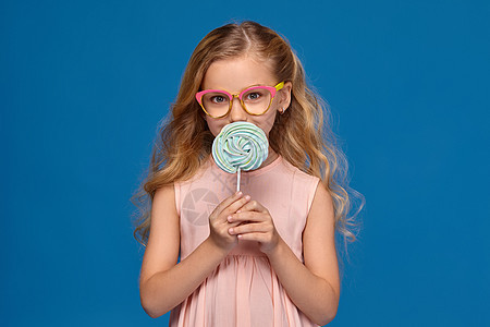 时尚的小女孩穿着粉红裙子和眼镜 手里拿着糖果 站在蓝色背景上 笑声配件金发手势头发孩子女孩耳朵皮肤青少年光学图片