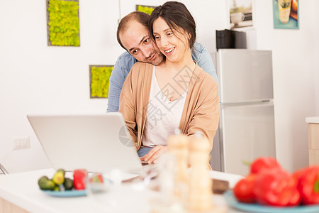 使用膝上型电脑的双亲微笑幸福男朋友工作厨房桌子成人丈夫女士蔬菜早餐图片