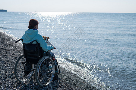 白种女人坐在轮椅上 和一只狗在海上海洋女孩拥抱卵石机动性压痛友谊海滩药品宠物图片