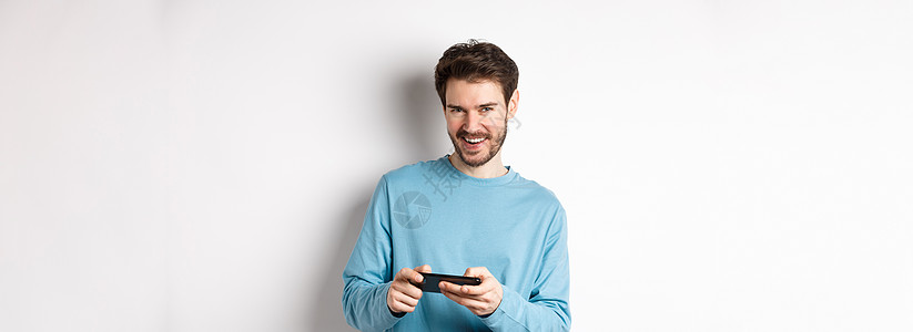 英俊的年轻人玩移动电子游戏 水平持智能手机 在摄影机喜悦下微笑 站在白色背景上站立商业促销胡子广告工作室发型办公室成功情绪技术图片