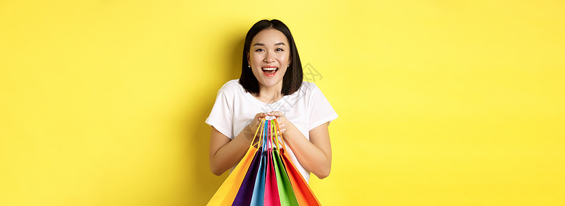 欢乐的亚裔女性直升机微笑并持有多彩购物袋 站在黄色背景下站立店铺促销学生发型礼物黑发购物者交易青少年女孩图片