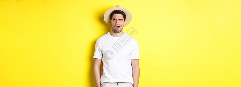 旅游和夏季的概念 戴着草帽的迷茫男旅行者 站在黄色背景上 看起来很困惑 什么也听不懂促销成人帽子广告技术男性商业工作室潮人胡须图片