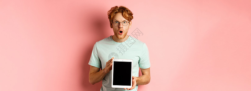 兴奋的年轻人留着红头发和胡须 查看在线促销 展示数字平板电脑屏幕 惊讶地盯着相机 站在粉红色背景上成人学生药片工作室红色发型眼镜图片